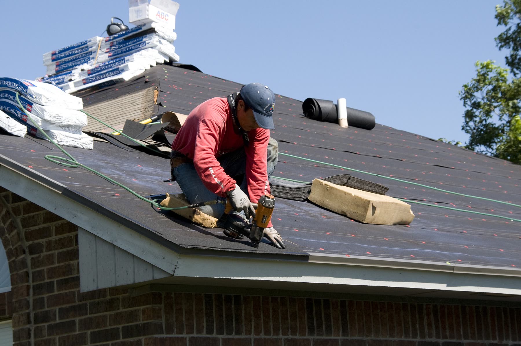 A roofer kneeling on a half-shingled roof, both hands on a nail gun